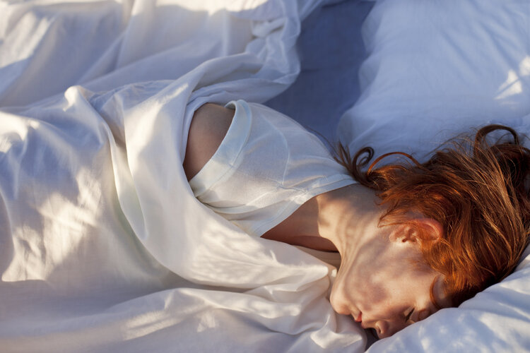 Woman with red hair sleeping in a bed with white sheets and a thin white shirt in soft, diffused natural light; portrait orientation.