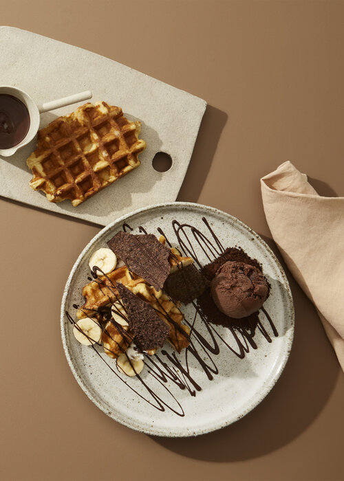 Overhead shot of waffle desserts with chocolate, bananas, and ice cream on a plate and serving board against a brown background.