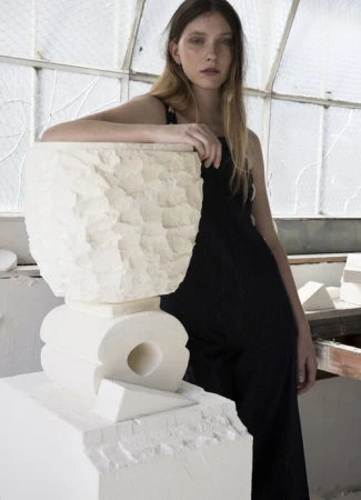 Young woman in a black dress leaning on a large carved white stone sculpture in a studio