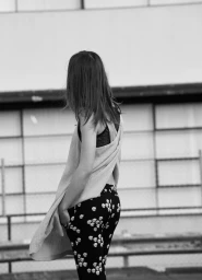 Woman with long dark hair seen from behind wearing patterned trousers and a draped top against an urban architectural background