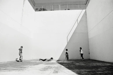 Four children playing in a large concrete courtyard with high walls under stark sunlight and deep shadows.