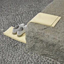 White leather shoes resting on pale yellow fabric next to a large grey stone on a bed of smooth river pebbles