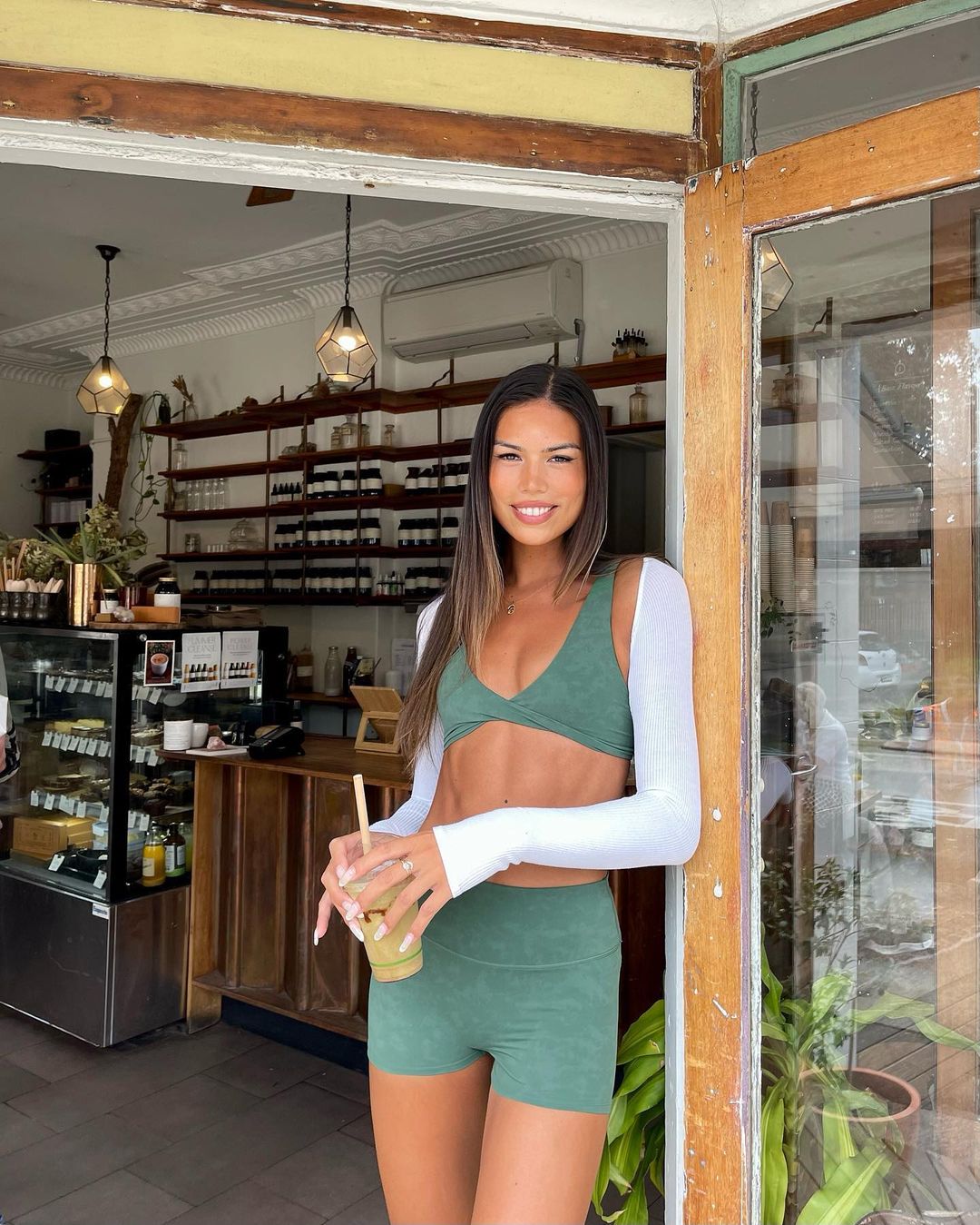 Adult woman in athletic wear stands in doorway, holding a beverage. Shelves and counter with supplies visible in background.