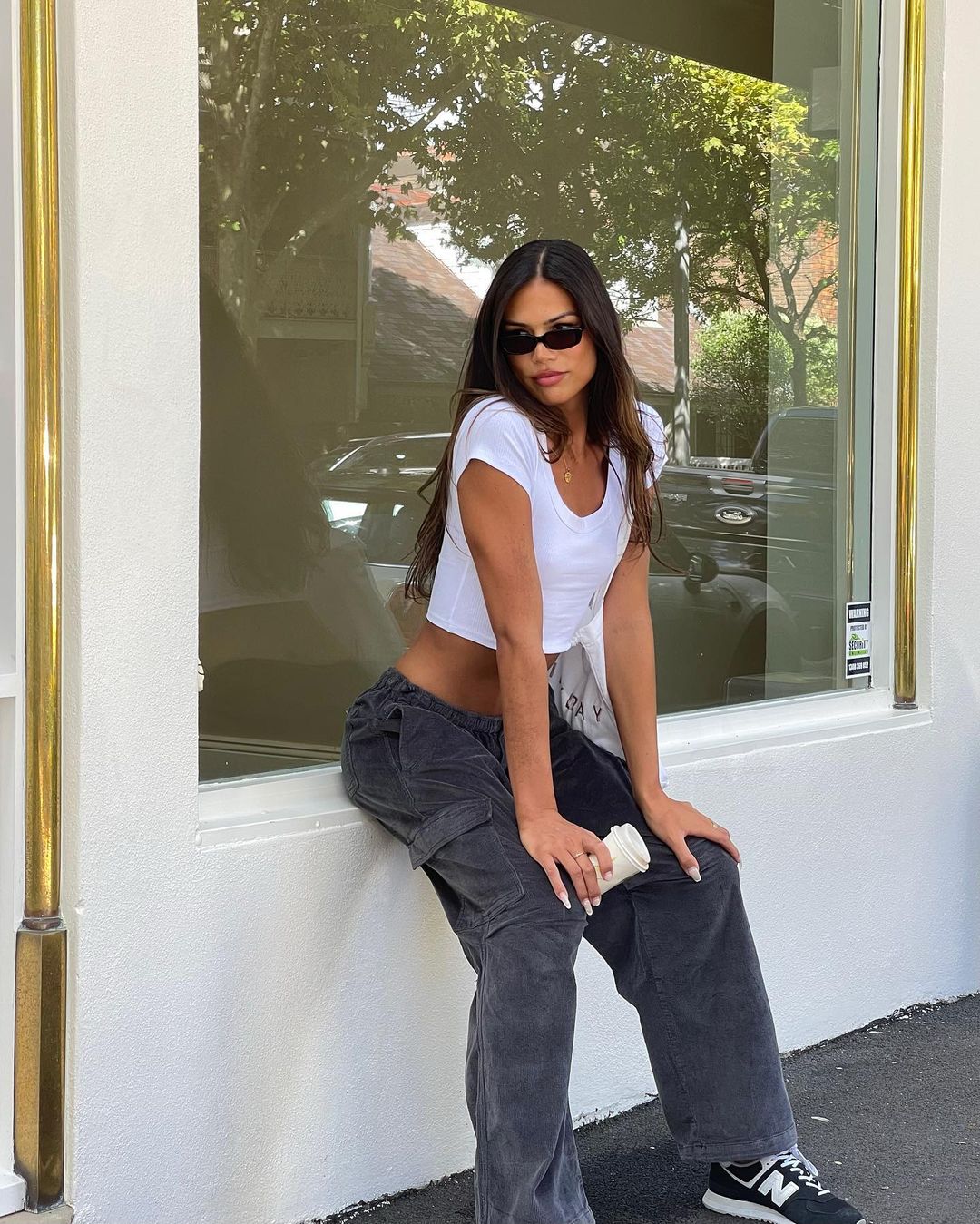 A brunette woman in a white top and grey pants poses seated outdoors with a building and trees in the background, holding a drink.