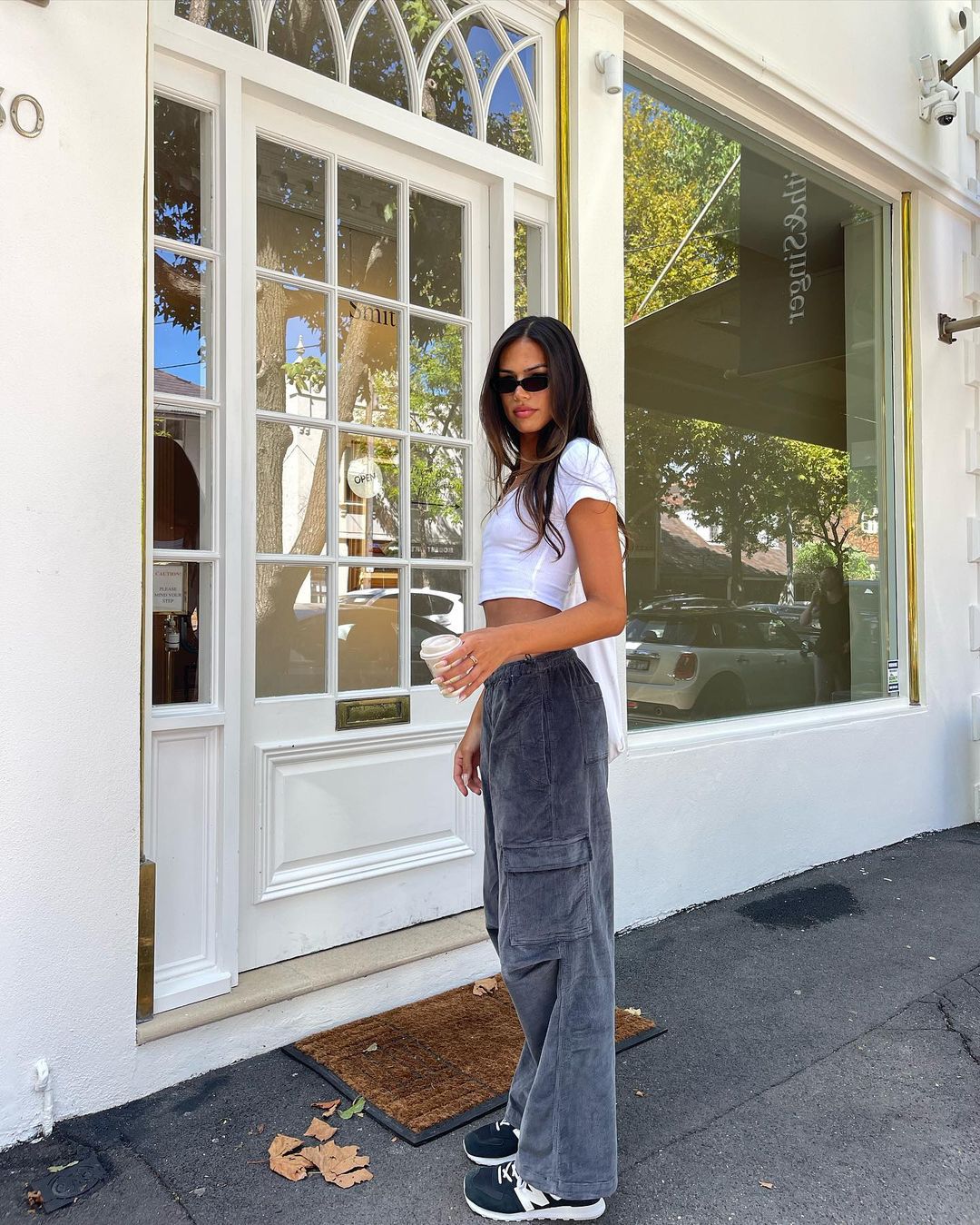 A young woman wearing sunglasses, a white top, and cargo pants stands outside a building with a white door while holding a cup.