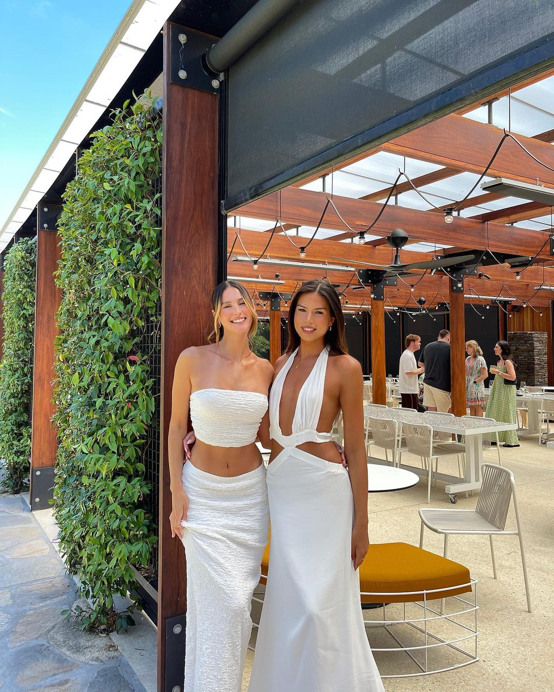 Two women in white dresses pose outdoors; one in a tube top and long skirt, the other in a halter dress with an outdoor event backdrop.