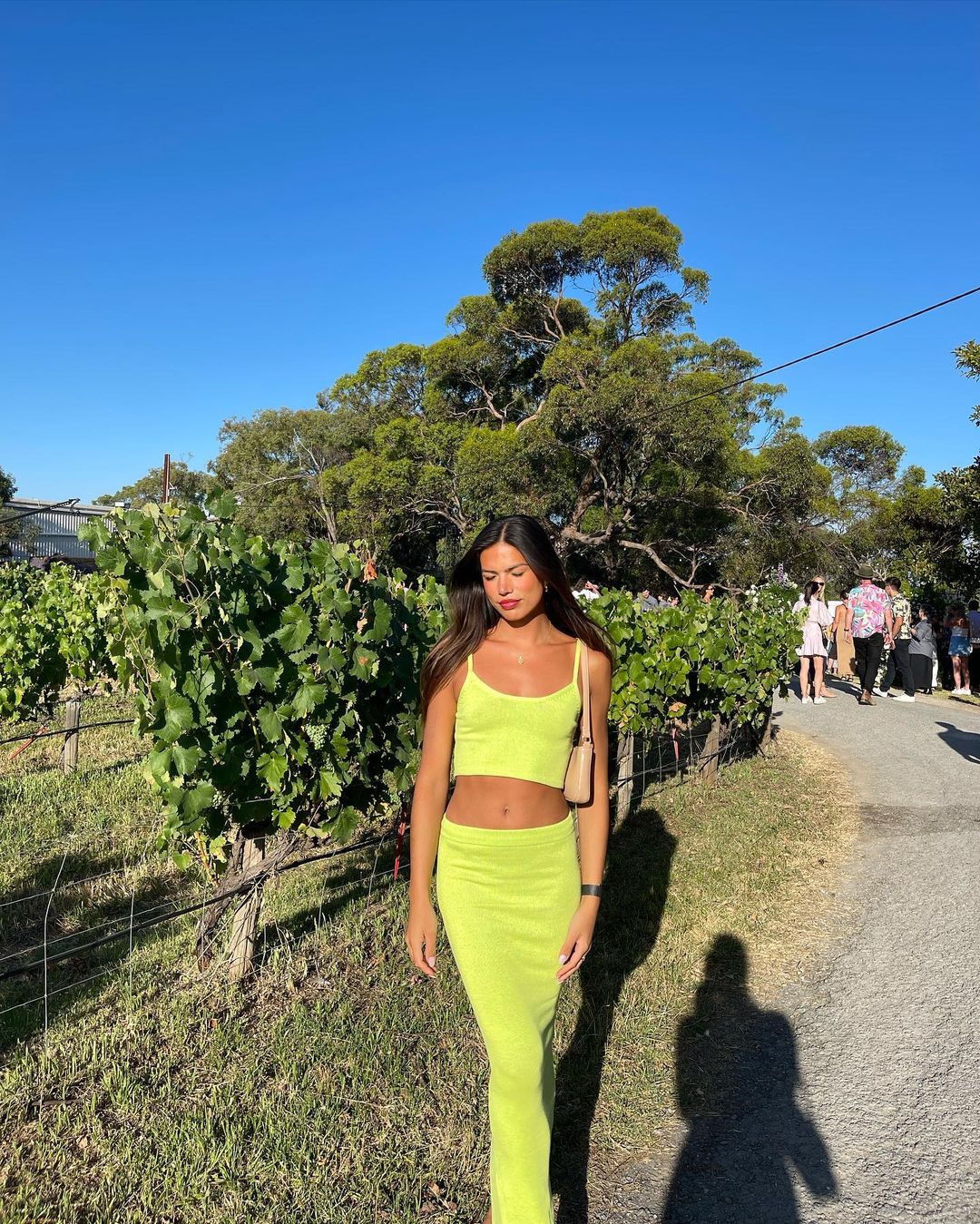 A woman in a matching neon yellow crop top and skirt stands in a vineyard on a sunny day, accessorized with a small shoulder bag.