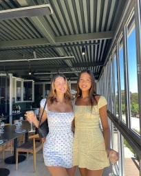 Two smiling women holding champagne glasses inside a restaurant with glass walls and vineyard views outside.