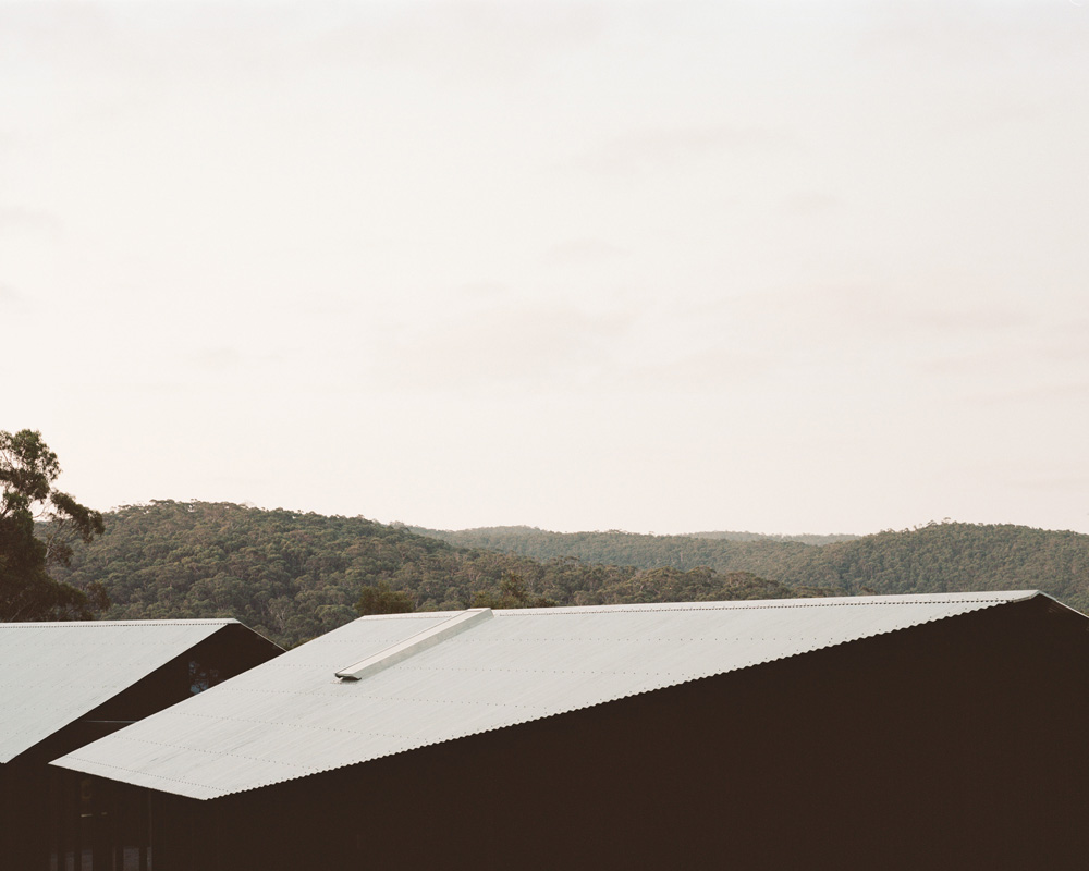 Architectural shot of the roofs of dark buildings topped with white metal under a bright sky with a wooded, hilly landscape in the distance.