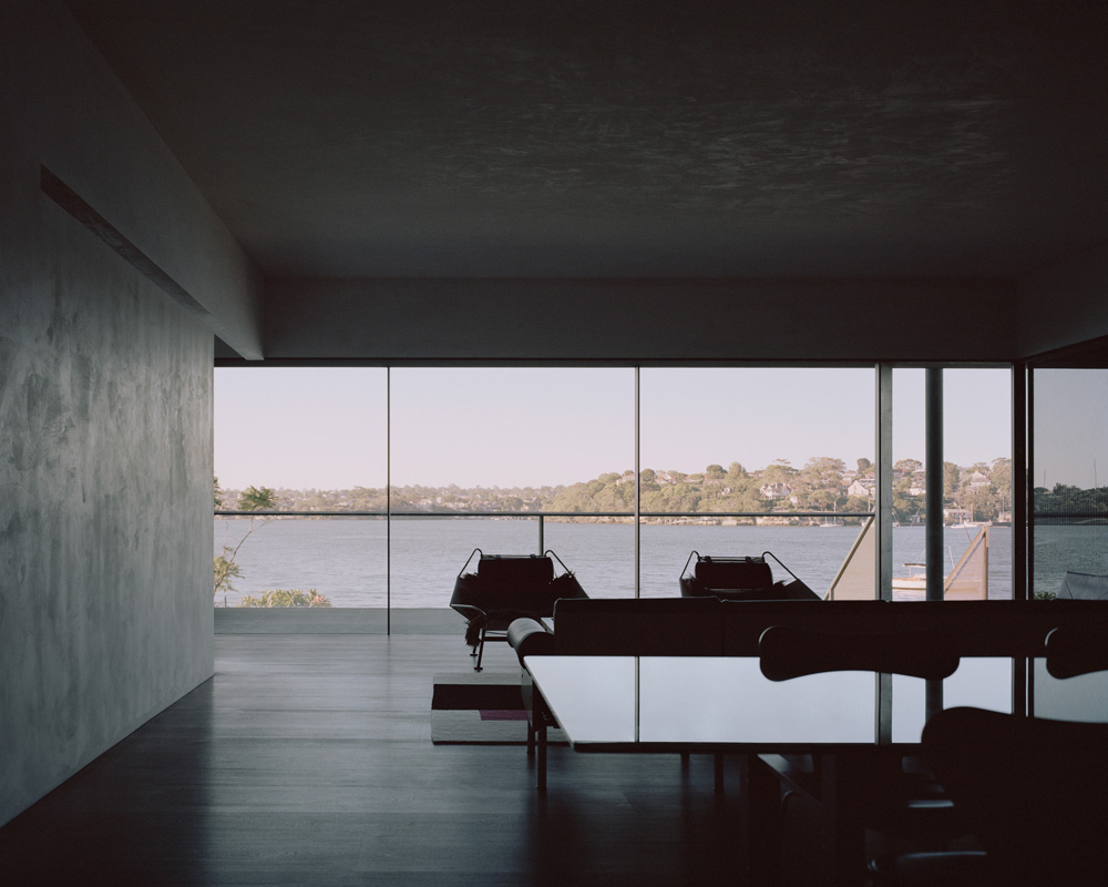 Modern interior with dark wooden floors, a long white table, and a wall of windows overlooking a river and trees in the background.
