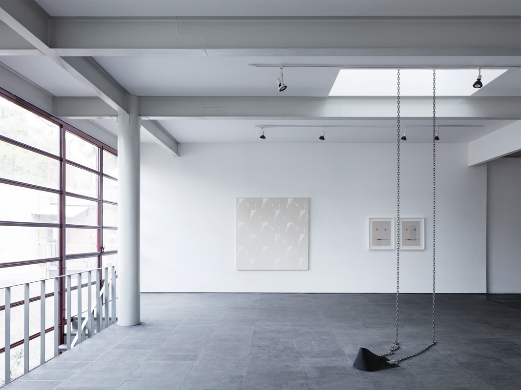 Minimalist art gallery interior with a black swing suspended by chains, artwork on white walls, and gray concrete floors.