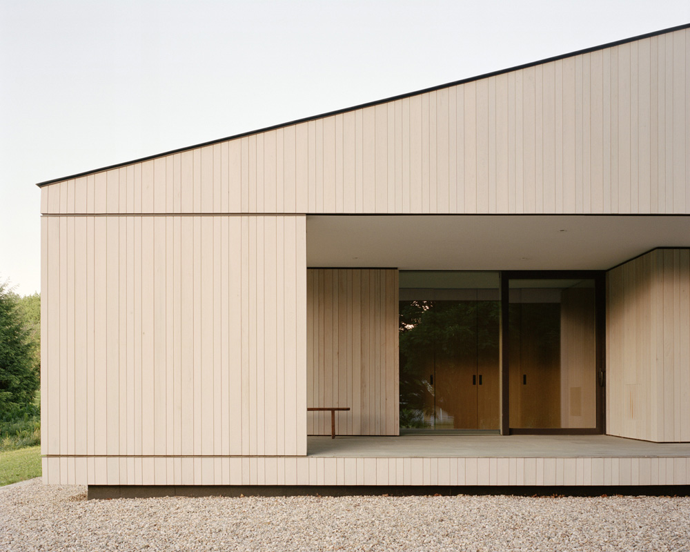 Exterior of modern home with light wood siding, minimalist design, gravel ground cover, and sliding glass doors in natural light.