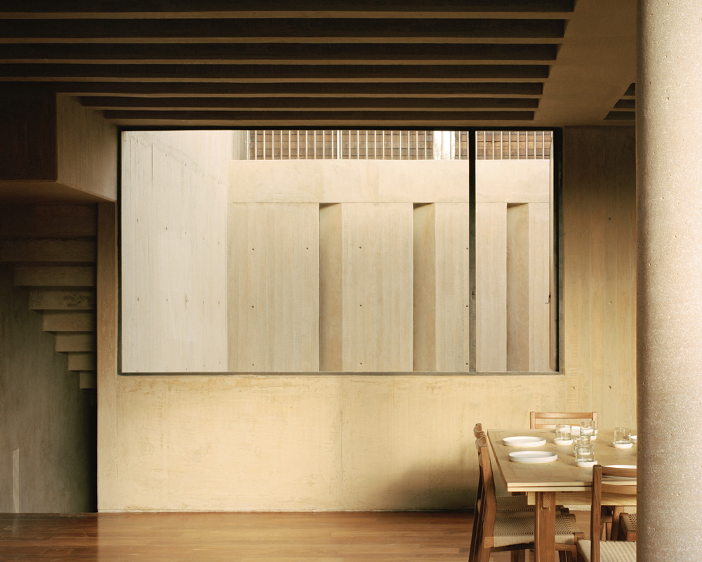Minimalist interior featuring a wooden dining table with tableware, a concrete column, and a large window showcasing an exterior wall.