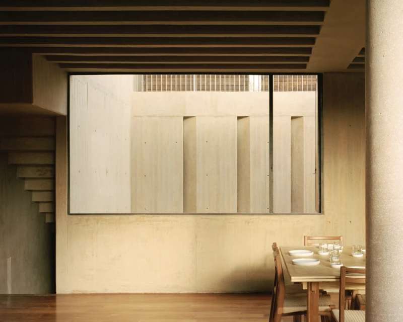 Minimalist interior featuring a wooden dining table with tableware, a concrete column, and a large window showcasing an exterior wall.