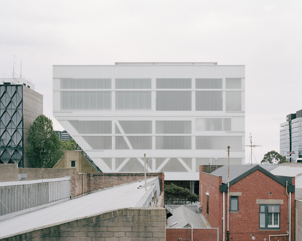 A contemporary white building with geometric design elements and horizontal window slats rises above low-rise brick structures in an urban setting.