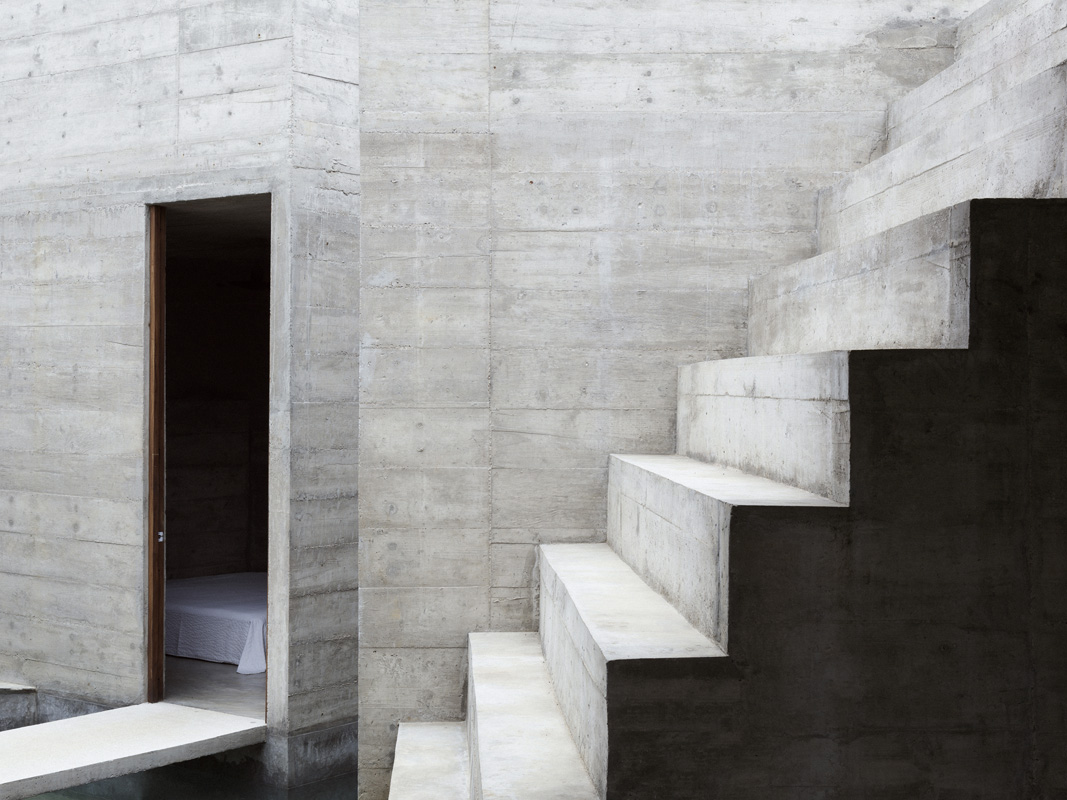 Architectural interior featuring concrete stairs and walls, with a doorway leading to a room with a bed, minimalist design.