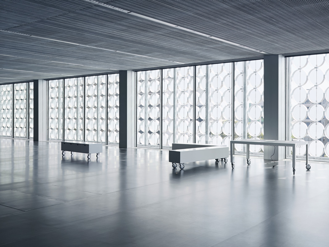 Large, modern interior with benches and tables on wheels, set against a backdrop of decorative windows and a paneled ceiling.