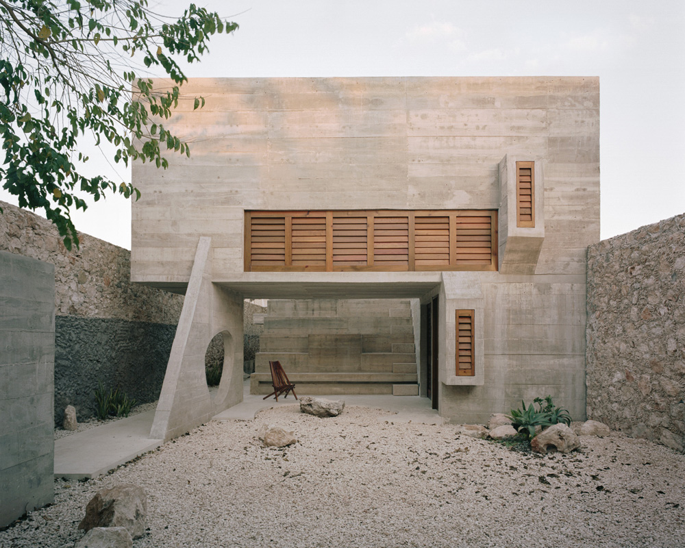 Exterior of a modern concrete house with wooden shutters and a gravel courtyard, showcasing minimalist architecture and natural light.