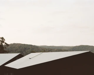 Corrugated metal roofs of buildings against a backdrop of distant wooded hills under an overcast sky, photographed by Rory Gardiner.