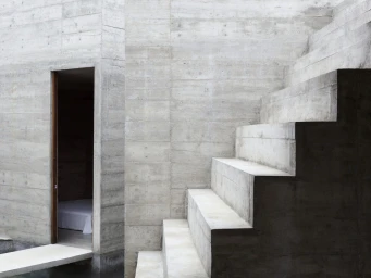 Exposed board-formed concrete staircase with block steps ascending beside a doorway revealing a lit interior room.