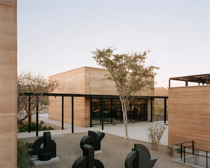 Architectural exterior photography showing a modern rammed earth building with black metal pergolas and abstract dark sculptures in a gravel courtyard.