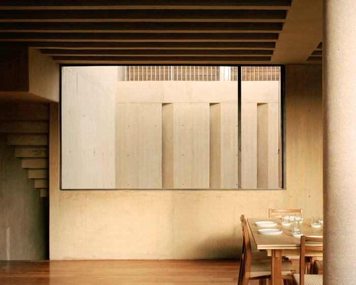 Interior architectural view looking out a large rectangular window onto a concrete courtyard wall, with wooden dining table and chairs in the foreground