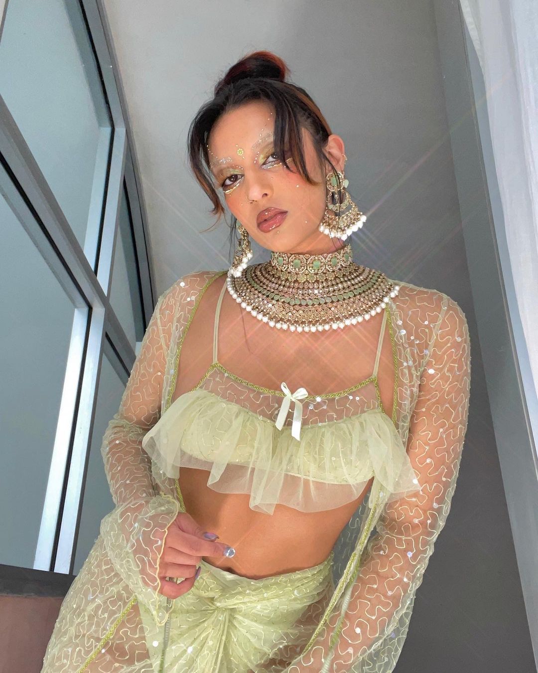 Woman with sparkling makeup in an embellished light green dress and jeweled necklace poses indoors, looking directly at the viewer.