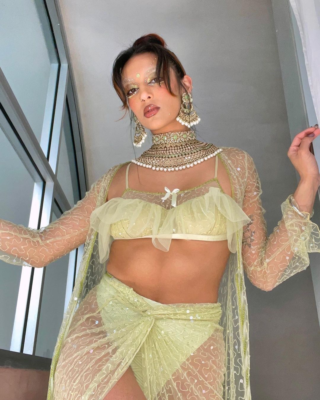 Woman in yellow lace lingerie, ornate jewelry, and face gems poses indoors near a window, showcasing a unique fashion style.