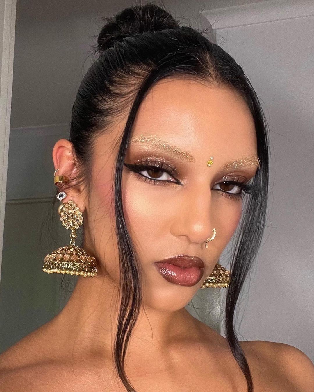 Close-up of a woman with dark hair in a bun, wearing eye makeup and statement earrings, posing indoors against a neutral background.