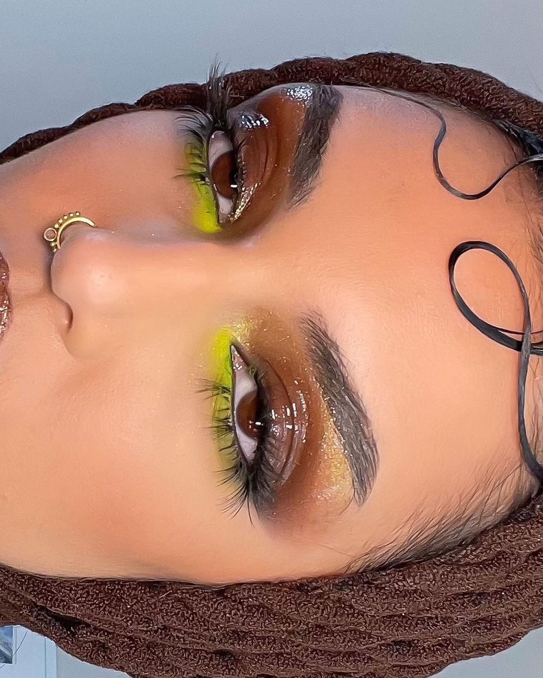 Eye-level portrait of a woman with brown eyes, fair skin, yellow eyeshadow, and a decorative nose ring, and a brown head wrap.
