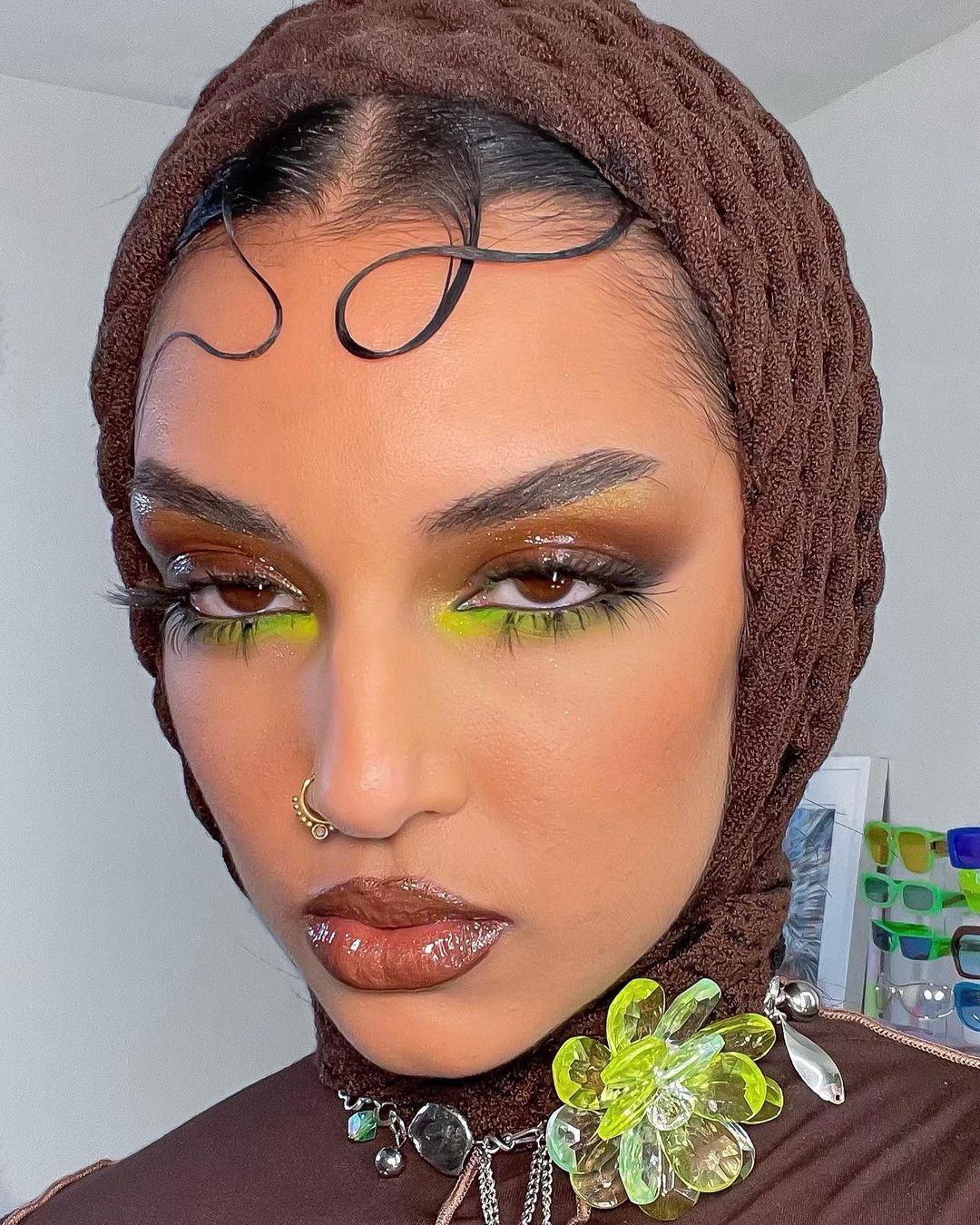 Close-up of a woman with brown headscarf, artistic eye makeup, glossy lips, nose ring, and a beaded flower pendant.