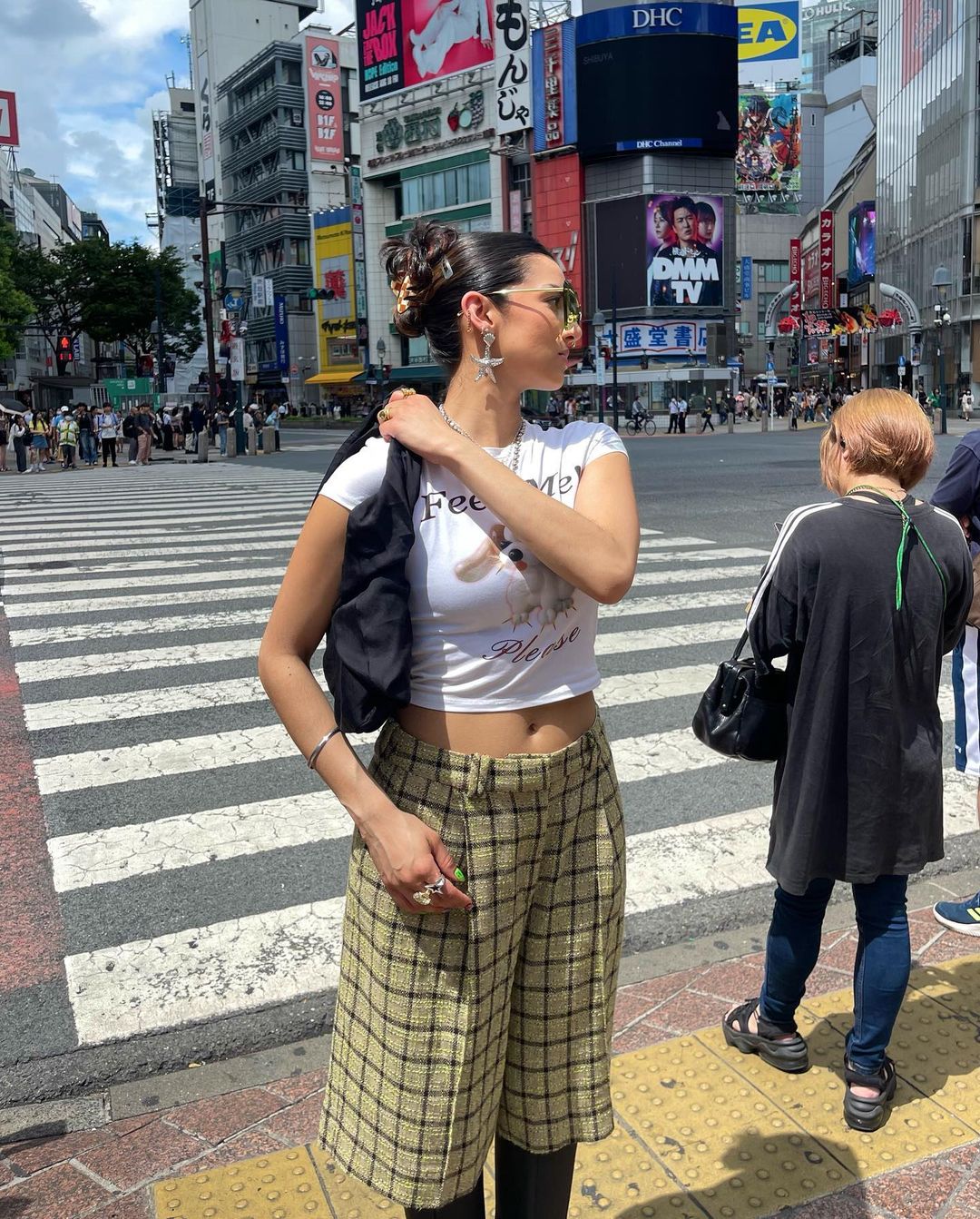 Woman in culottes stands on crosswalk in Shibuya, Japan, with buildings and billboards in the background, on a sunny day.