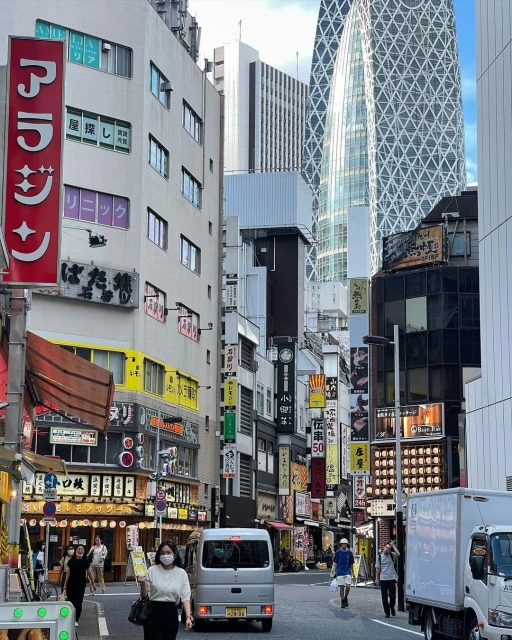 City street view in Tokyo, Japan, showcasing urban architecture, commercial signage, pedestrians, and vehicular traffic on a cloudy day.
