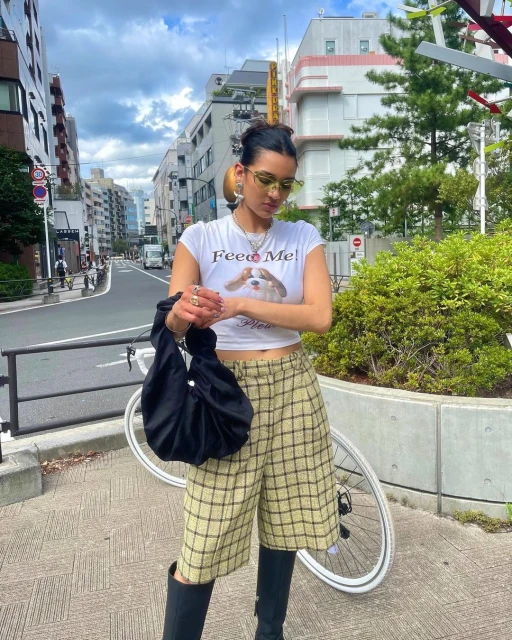 Woman in casual street style poses in an urban setting with buildings, wearing a graphic tee, plaid shorts, and knee-high boots.