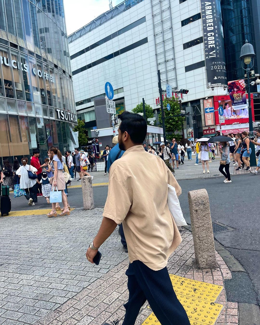 Man in casual attire walks across a brick-paved city street in Tokyo, with a crowd and modern buildings visible in the background.