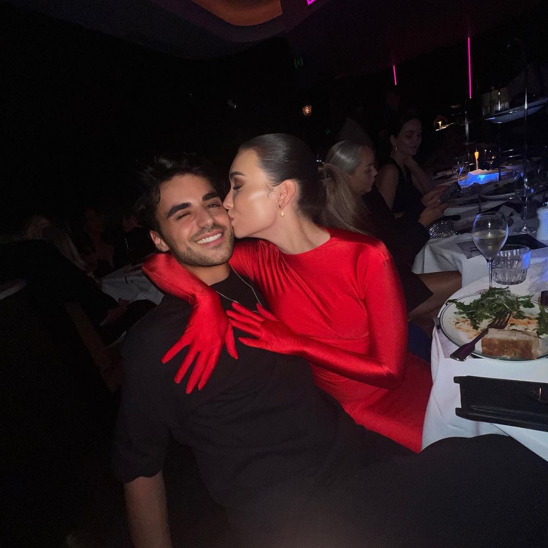 Couple in formal attire at an event; the woman kisses the smiling man on the cheek, both are indoors with a party atmosphere.