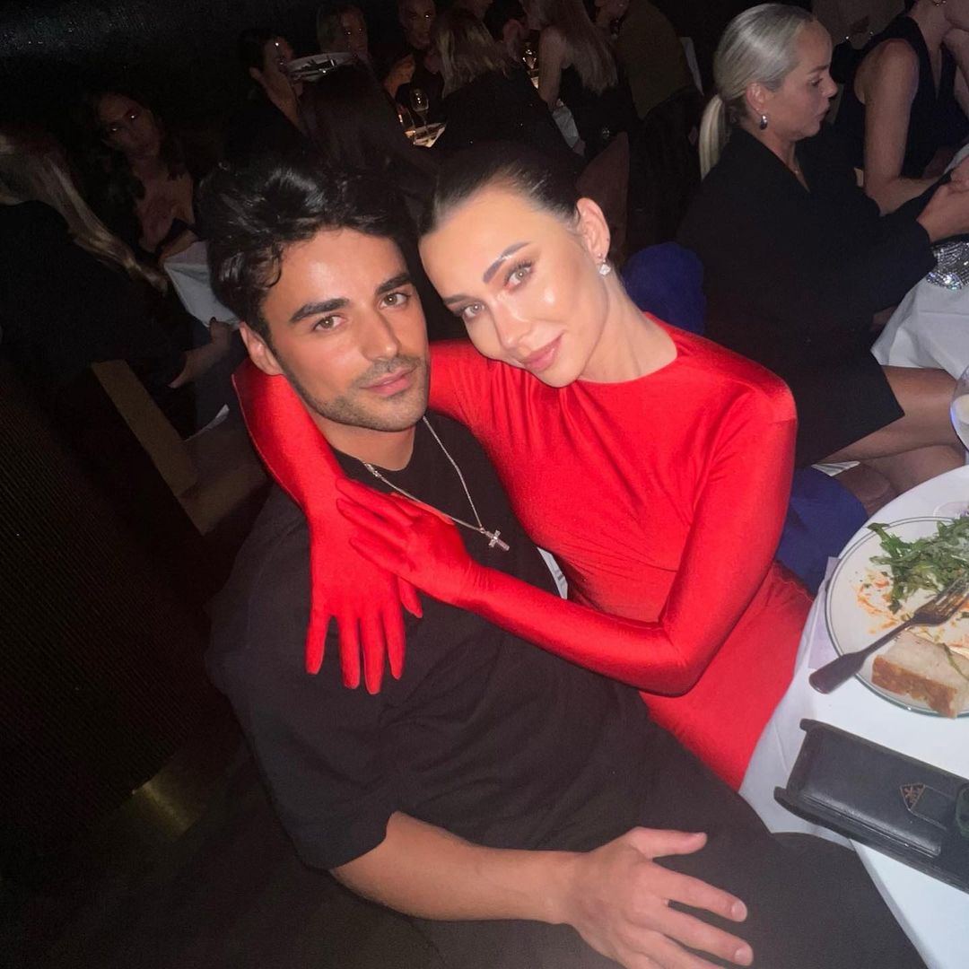Woman in red dress and gloves embraces man at a table indoors, with background of seated people at tables, creating an intimate gathering.