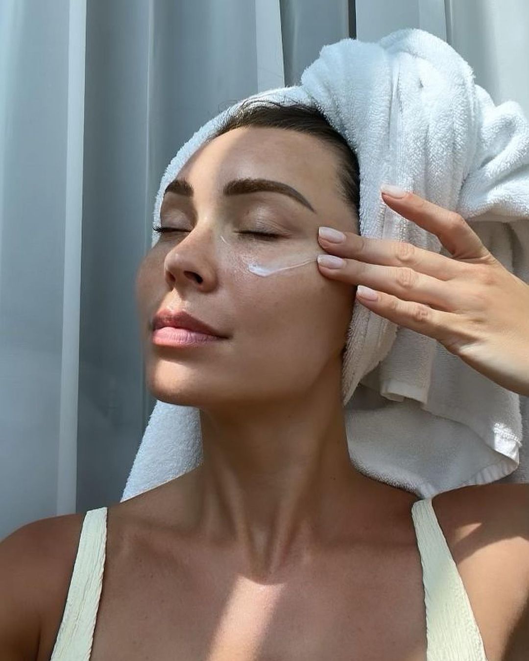 Close-up of a woman applying cream under her eyes with a towel wrapped around her head in a bright indoor setting.