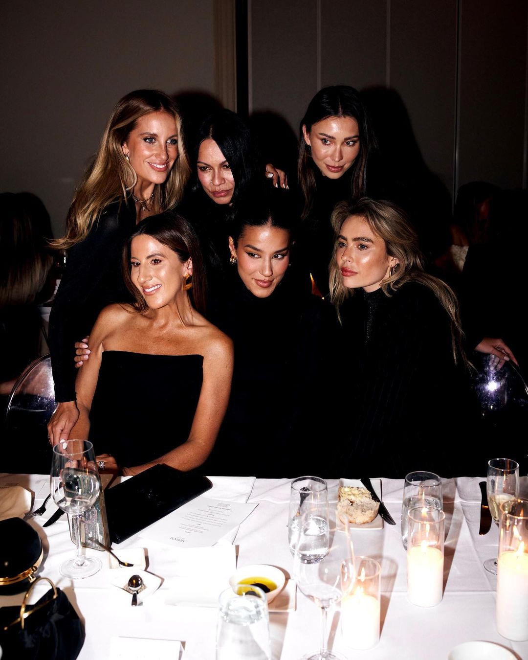 Group of six adult women posing at an indoor event with table setting, wearing black clothing, fair skin tones, and light makeup.