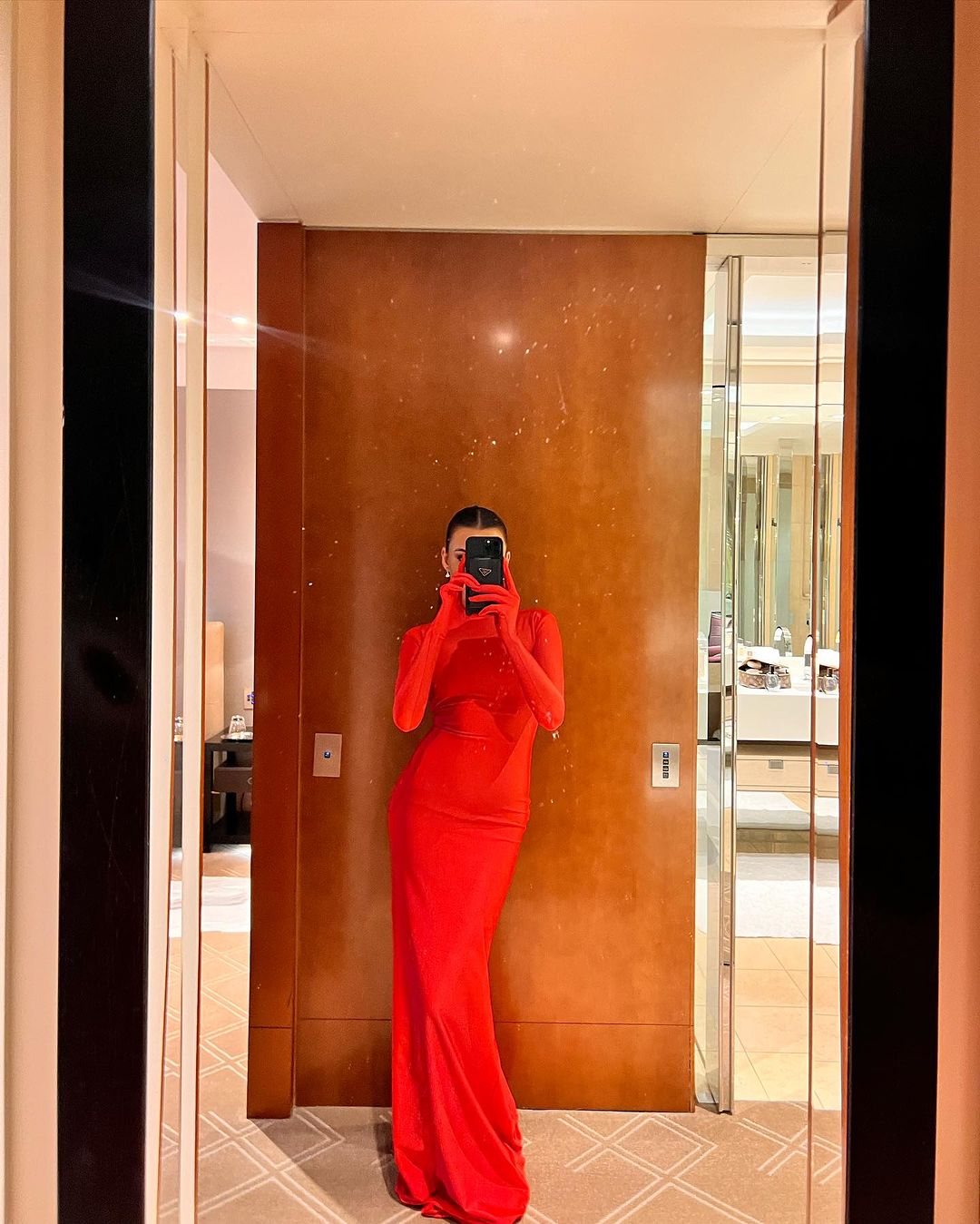 Woman in a long red dress takes a mirror selfie in a neutrally toned room with partial views of furniture and bathroom amenities.
