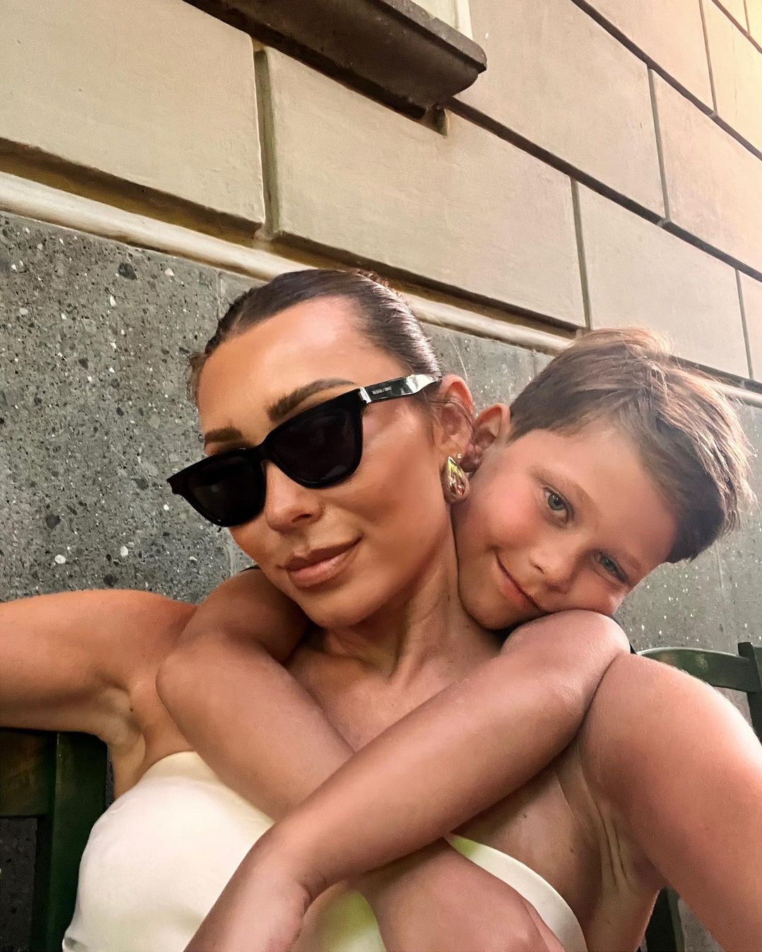 Mother and child embrace outdoors. Woman wears sunglasses, off-shoulder top, and earrings. Boy hugs her with a smile in the frame.