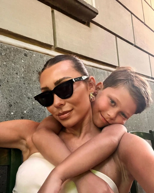 Mother and child embrace outdoors. Woman wears sunglasses, off-shoulder top, and earrings. Boy hugs her with a smile in the frame.