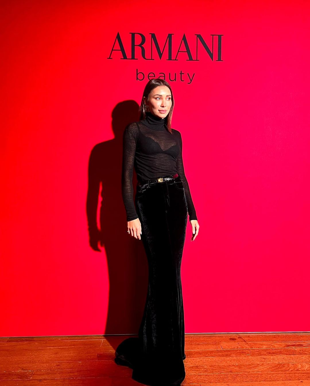Elegant woman in black turtleneck and velvet skirt poses against a red backdrop with the Armani Beauty logo, showcasing a beauty campaign.
