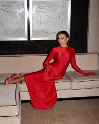 Woman in a long red dress with matching gloves and heels seated on a beige sofa in a modern interior.
