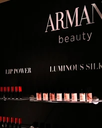 Armani beauty display with Lip Power and Luminous Silk products on illuminated glass shelves against a black wall.