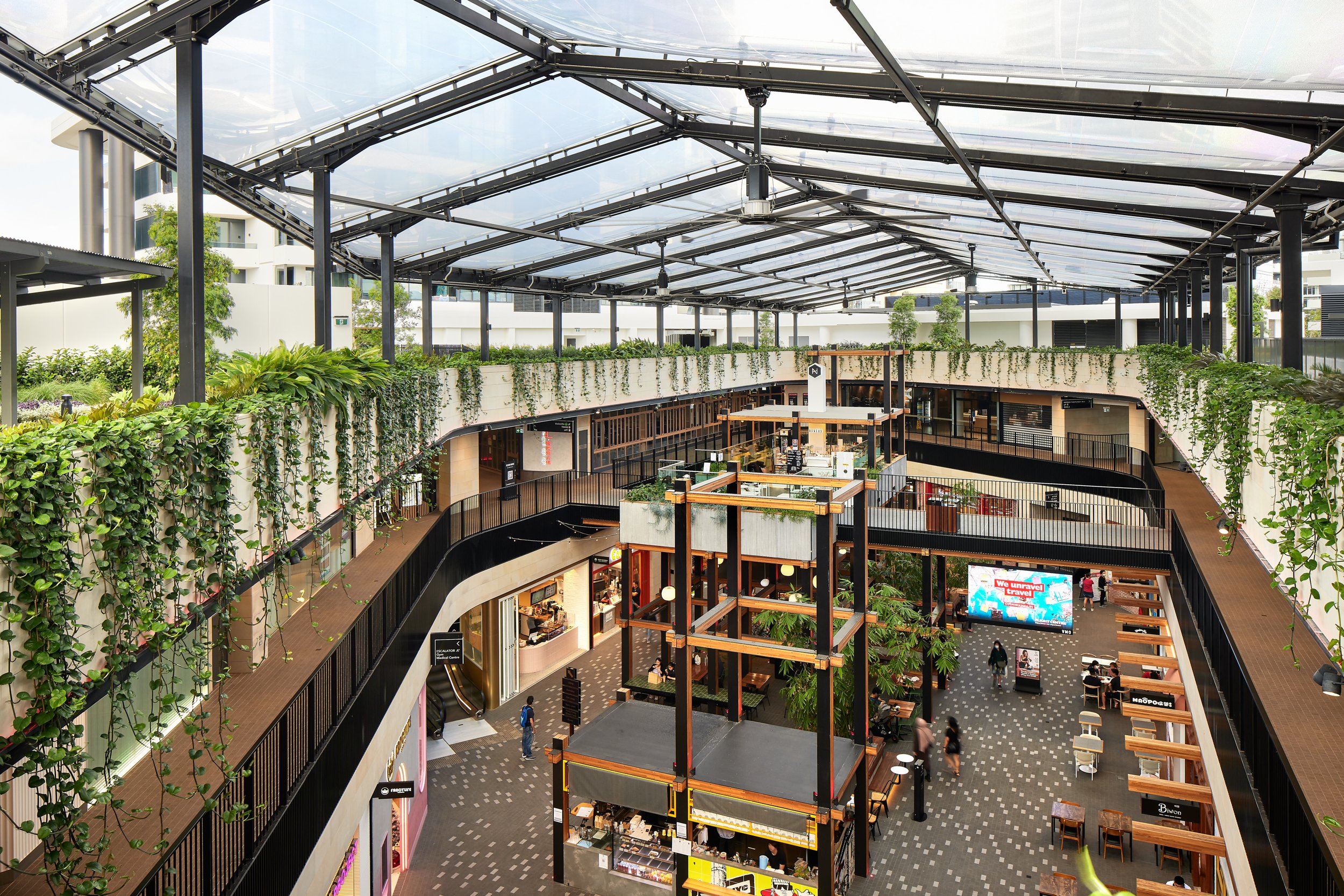 Multi-level indoor shopping mall with open-air roof, greenery, visible shops, tables, and people walking and sitting. Bright daytime.