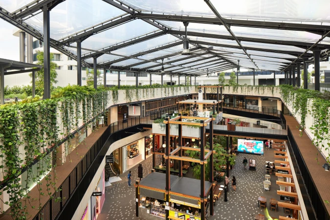 Multi-level indoor shopping mall with open-air roof, greenery, visible shops, tables, and people walking and sitting. Bright daytime.