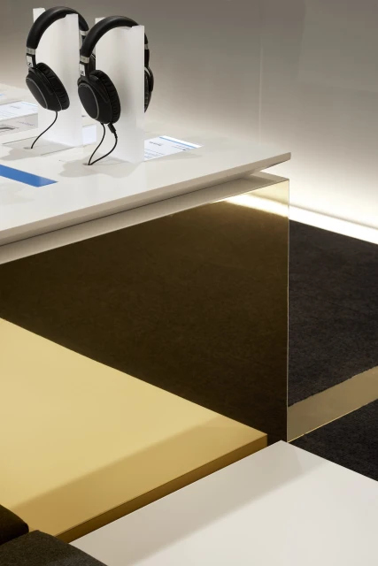 Two black over-ear headphones are displayed on a white table with geometric color-blocked panels in white, gold, and black.