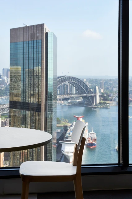 Interior view of an office with a round white table and chair overlooking the Sydney Harbour Bridge, buildings, and blue water.