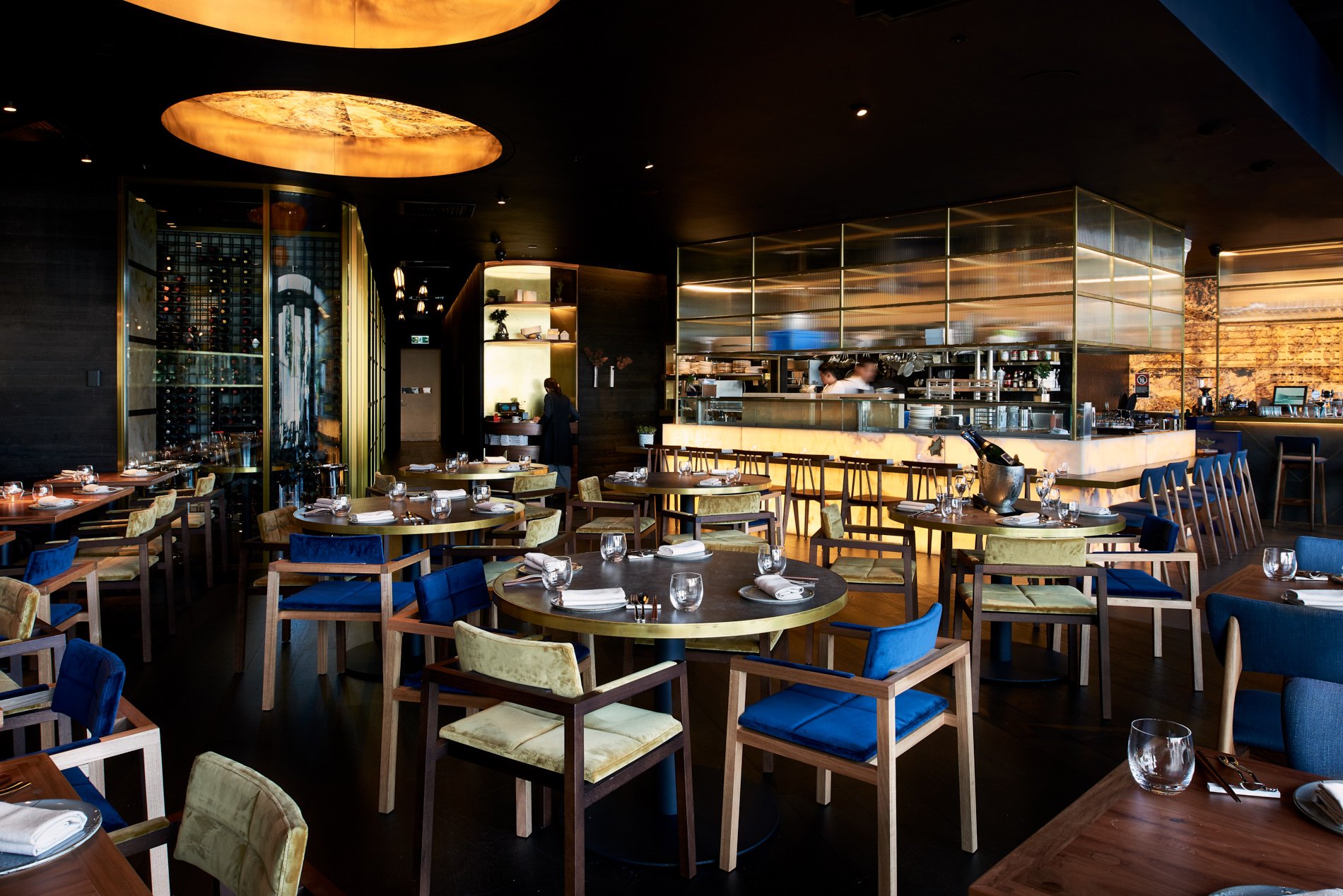 Upscale restaurant interior showcasing set tables with blue and yellow chairs, soft lighting, and bar area in the background.